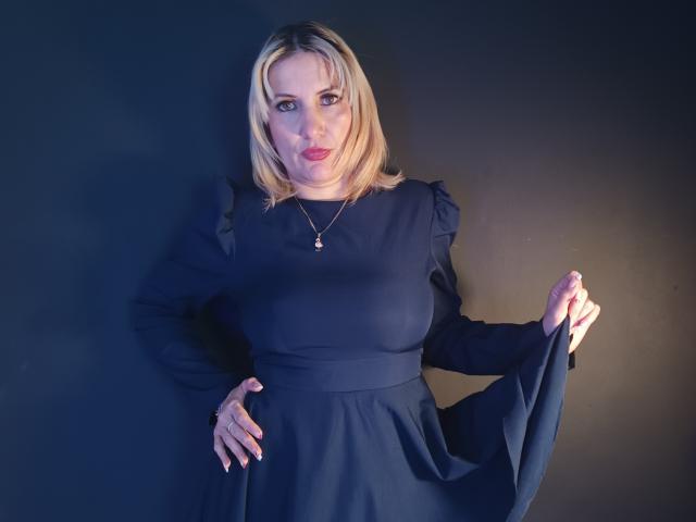 GiuliaAddams, avec des cheveux blonds, portant une robe noire, les mains sur les côtés, une main sur la hanche, rouge à lèvres, collier, fond sombre, debout, souriant légèrement, seins visibles, corpulence moyenne..