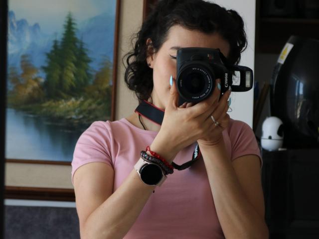 GabrielaMoore with curly black hair, wearing a pink shirt, holding a black camera, covering her face, wearing a black strap, red bracelet, and a wristwatch, in a room.