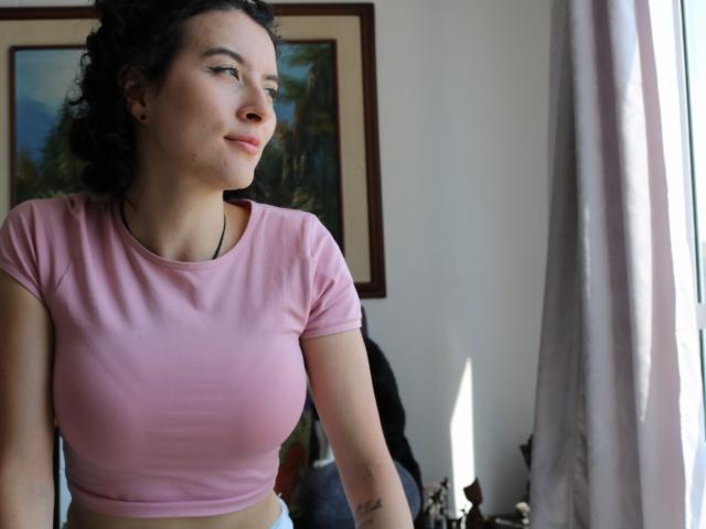 GabrielaMoore with curly dark hair, pink crop top, showing her small tits, standing by a window, smiling slightly, natural light, white curtains, painting in background.