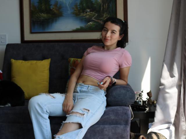GabrielaMoore with small tits, wearing a pink crop top and ripped white jeans, sits on a gray couch, smiling, with dark curly hair, and a black cat nearby.