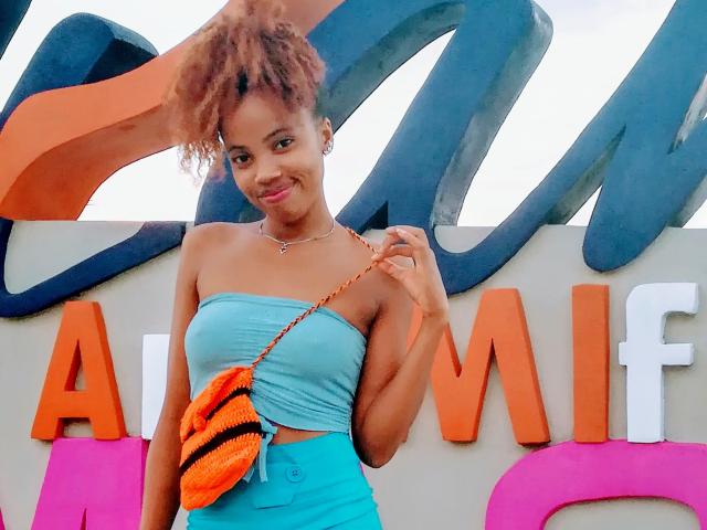 HotEllySexy with light brown skin, curly hair, small tits, wearing a turquoise strapless top, orange and blue bag, and blue pants, smiling at the camera.