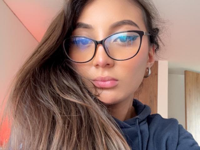 DianaFernandezz with blue-tinted glasses, pink lips, and long brown hair, wearing a black hoodie, showing off her cute face and slightly parted lips.