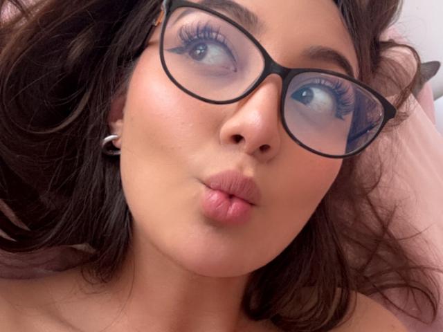 DianaFernandezz has pouty pink lips, big brown eyes, and wears black glasses. She's got wavy brown hair, and a cute nose. Her tits are not visible.