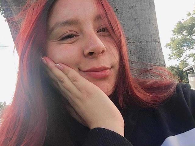 EmaJames, con cabello rojo, piel clara y labios rosados, sonríe suavemente, con la mano apoyada en su mejilla. Lleva una blusa negra y blanca y está de pie junto a un árbol..