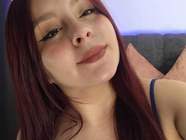 EmaJames met rood haar, lichte huid en blauwe eyeliner, die lichtjes glimlacht, waarbij haar volle lippen en kleine neus zichtbaar zijn. Ze draagt een blauwe band over haar bovenlichaam en heeft een subtiele blush op haar wangen..