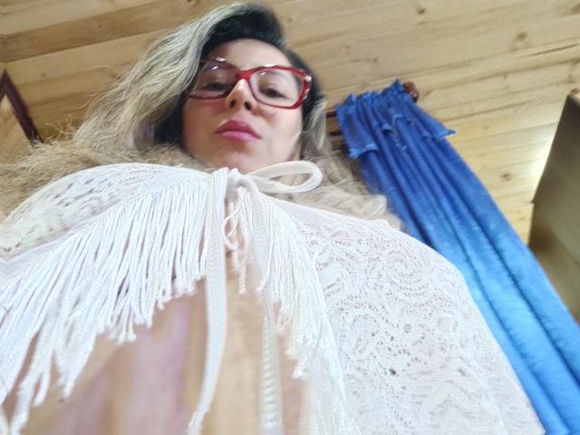 Contelaya with blonde hair, red glasses, and white lace top, showing her tits, looking down at the camera, blue curtain in the background. Wooden ceiling above.