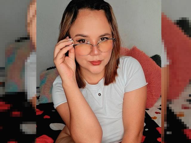 Natalie Rey con gli occhiali, che si tocca il naso, indossa una camicia bianca, è seduta con un sorriso appena accennato, ha i capelli castani, la pelle chiara, rossetto rosso, seno piccolo, luce soffusa sul viso..