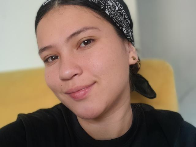 CelesteDash a un bandana noir, des yeux marron et un petit sourire. Elle porte une chemise noire avec des boucles d'oreilles dorées, et son visage présente une peau claire avec des taches de rousseur..