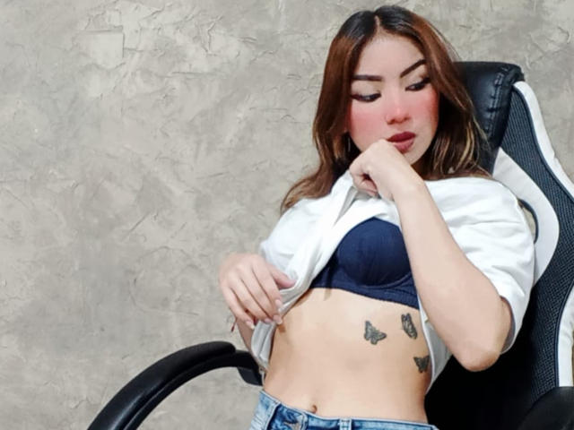 AshleyMiler with her tits exposed, lifting a white shirt to show a blue bra and butterfly tattoos on her flat stomach, sitting in a black chair.