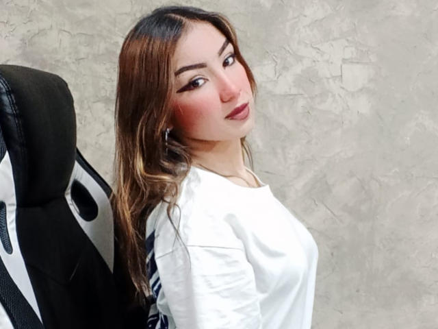 AshleyMiler has wavy brown hair, white shirt, and dark lipstick, sitting on a black and white gaming chair, looking over her shoulder with a subtle smile.