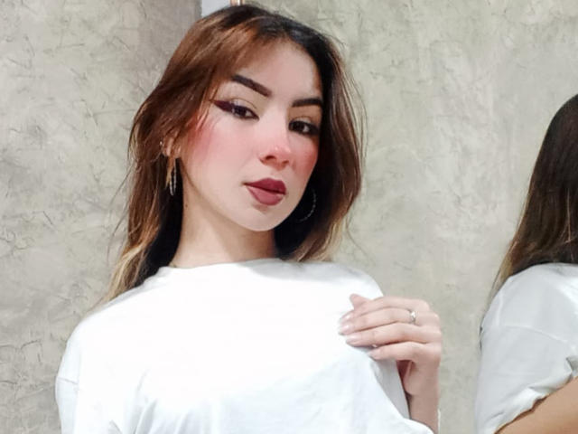 AshleyMiler with brown hair, pink blush, winged eyeliner, hoop earrings, and a white shirt, holding her shirt, showing a ring, and a slight pout.