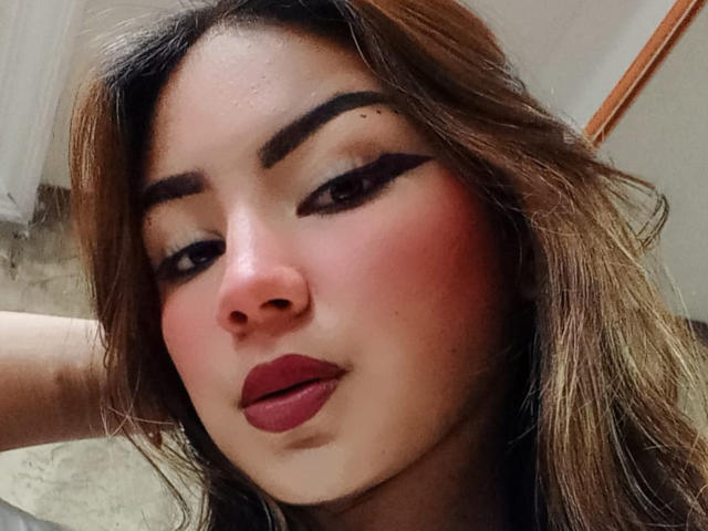 Ashley Miller tiene un rostro atractivo con cejas pobladas, delineador de ojos oscuro, labios rojos y una ligera sonrisa. Tiene el cabello castaño y húmedo, y un ligero rubor rosado en las mejillas..