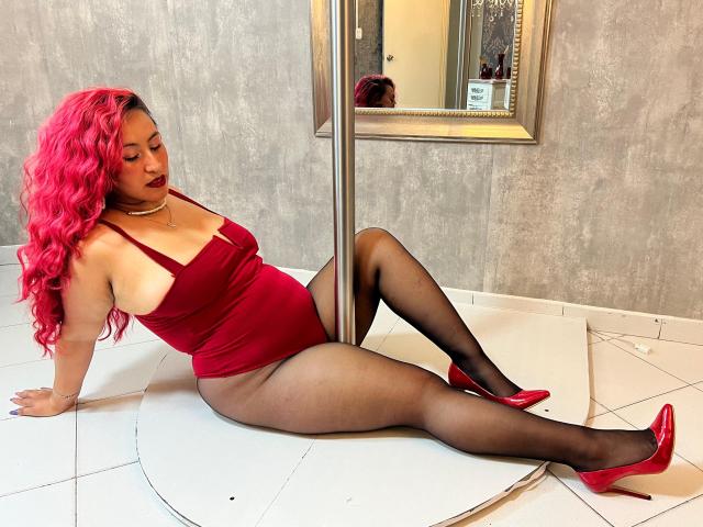 VictoriaTheas with bright pink curly hair, red velvet bodysuit, black pantyhose, and red heels, sitting on white tile floor, legs spread, leaning on silver pole, showing her ass.