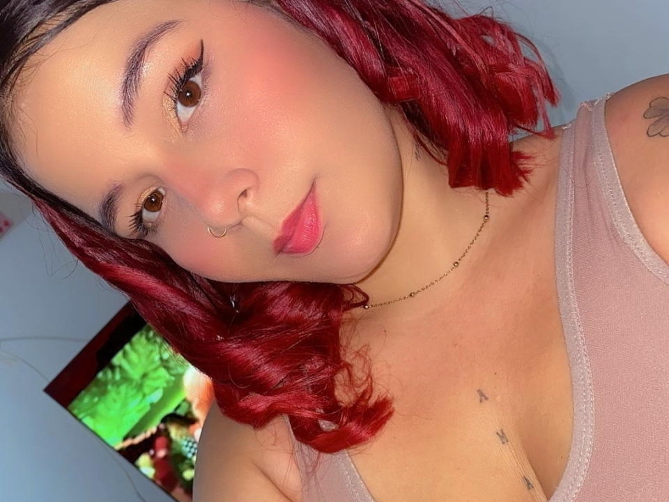 KattyMorganX with red hair, nose ring, and pink lips, wearing a pink tank top, showing small tits and a tattoo on her right shoulder. Her brown eyes are soft.