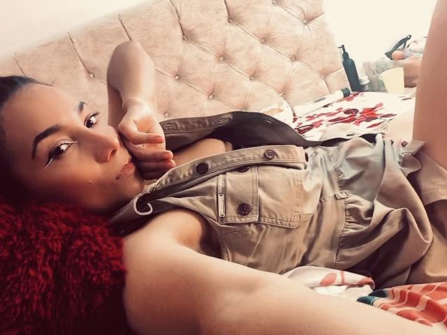 AmberDemons with red hair, lying on bed, wearing a tan sleeveless dress, showing small tits, black bra, and a lip ring, making a peace sign, smiling.