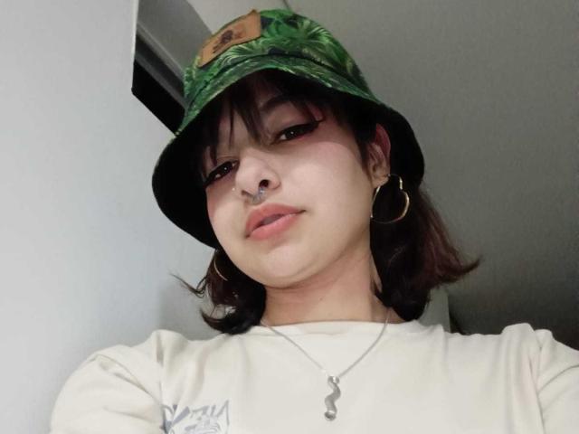 LaraAhB porte un chapeau seau vert, un anneau de nez, des boucles d'oreilles cerceaux et une chemise blanche avec un collier. Elle regarde l'appareil photo avec un léger sourire..
