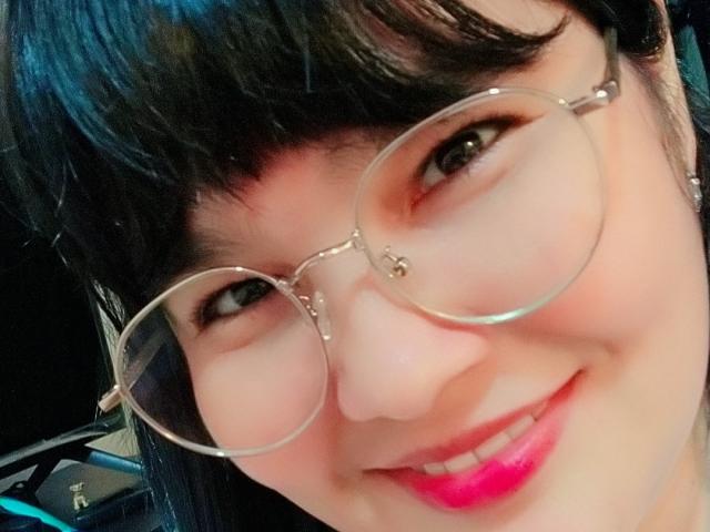 PrittyAlicia with black bangs, wearing round glasses, has shiny pink lipstick, and a cute smile, looking slightly to the side. She's got a small nose ring on her right.