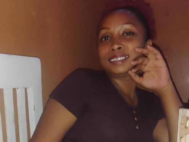 Cathionass with dark brown skin, medium-sized tits in a black shirt, red curly hair, smiling, hand touching cheek, white chair on left, brown wall in background.