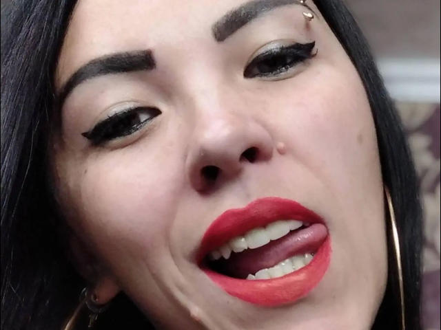 AmandaHot-hot's face with black eyeliner, red lipstick, and tongue out. She has a nose piercing, hoop earrings, and a facial mole. Her eyebrows are thick and dark.