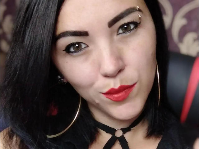 AmandaHot-hot has black hair, red lipstick, and a nose piercing. She's wearing a choker and large hoop earrings, with a slight smile. Her eyebrows are thick and arched.