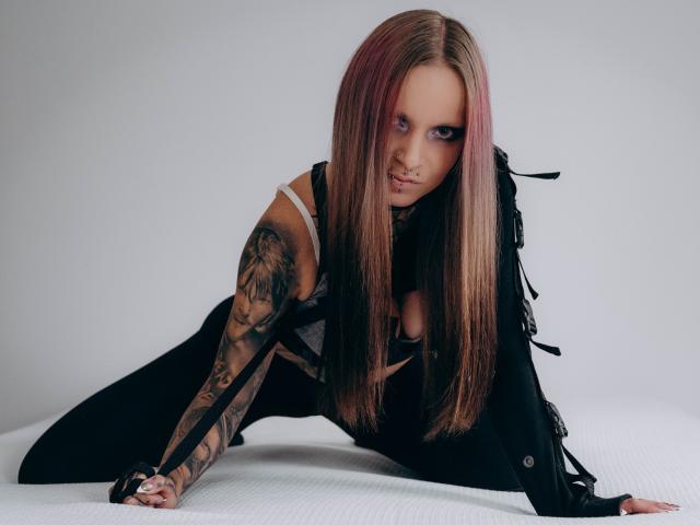 InesCreazy with long pink-and-brown hair, tattoos on her right arm, black fringed jacket, and white tank top, on all fours, looking at camera with a slightly open mouth.