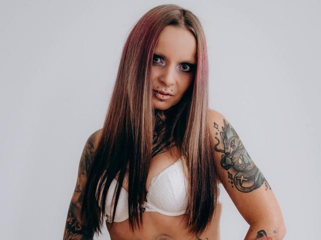 InesCreazy with long brown and pink-streaked hair, white lace bra, multiple tattoos, nose ring, and lip piercing, staring seductively at the camera. Boobs, tits, tattoos, piercings.