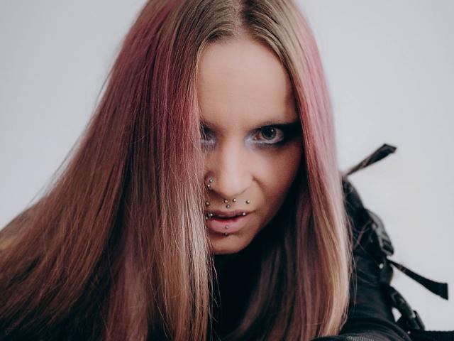 InesCreazy with pink and brown hair, nose piercings, lip piercings, and intense blue eyes, looking directly at the camera with a slightly angry or determined expression.