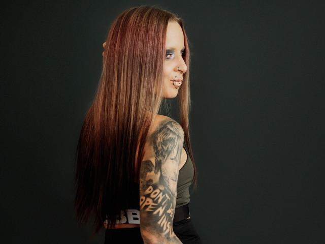 InesCreazy has long, straight, brown-red hair, tattoos on her right arm, and is looking over her shoulder with a small smile. She's wearing a green top and black pants.