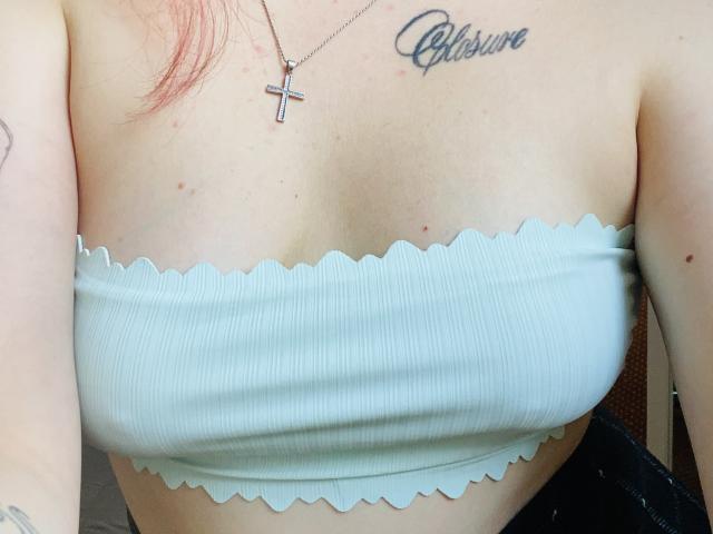 KarKutie's small tits in a scaggled-edge, ribbed, strapless top. She's got a cross necklace and "Blessure" tattoo above her right tit. Light skin with pinkish hair.
