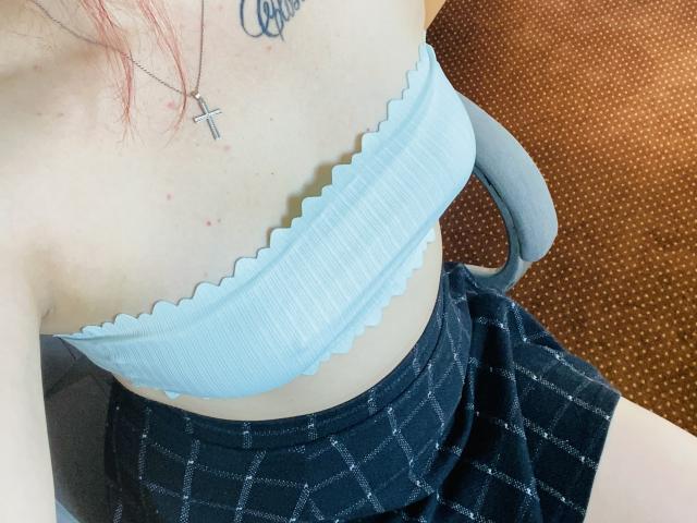 KarKutie with pale skin, small white ribbed tube top showing tits, black checkered skirt, cross necklace, pink hair, sitting in gray chair with orange patterned seat.