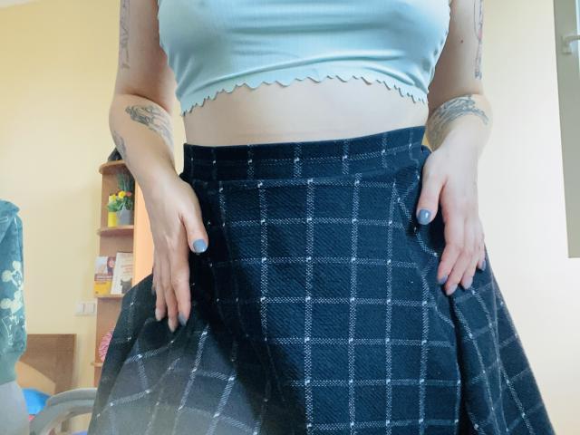 KarKutie wearing a light blue crop top with scalloped edges, showing her midriff, and a dark blue plaid skirt, hands on her skirt, tattoos on her arms.