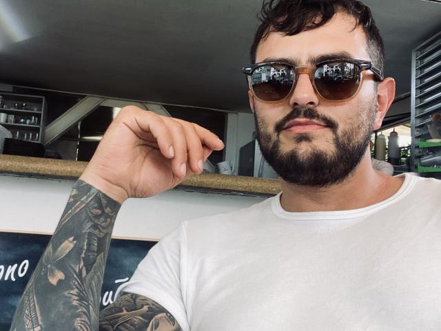SebastianBetacourt with a beard, wearing brown tortoiseshell sunglasses, white t-shirt, and tattooed arm, raising his right hand in a casual, confident selfie.