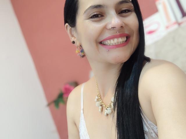 CharlotteCrofort with black hair, nose and lip piercings, smiling, wearing a white lace top, gold necklace, colorful earrings, showing her tits slightly, in a bright photo.
