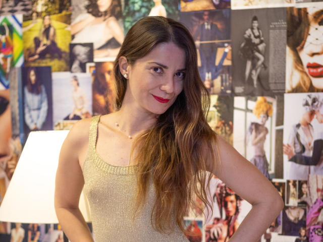 NatashaKute with long brown hair, red lipstick, and a gold glitter tank top, standing with hands on hips, smiling at the camera, surrounded by magazine photos.