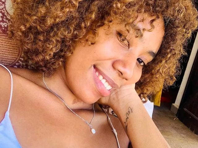Kenjya a les cheveux bruns bouclés, elle sourit en montrant ses dents blanches, et elle porte un collier en argent. Elle repose sa tête sur sa main, révélant un petit tatouage sur son bras droit..
