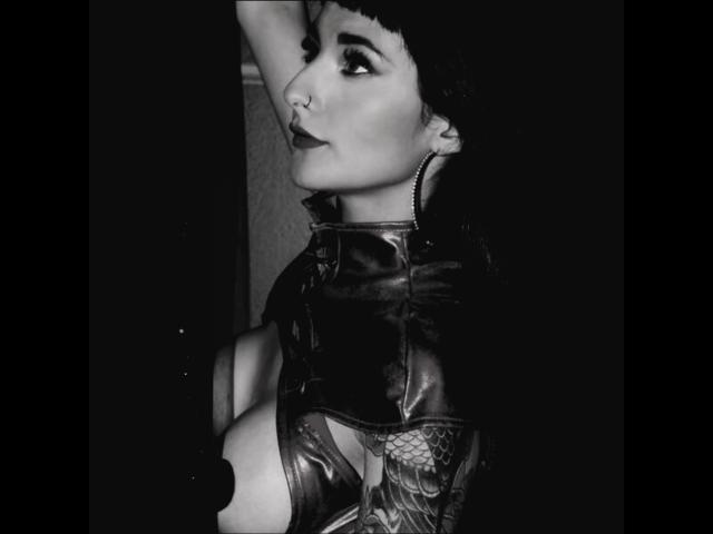 Carll has dark makeup, a nose ring, and wears a black leather outfit with cutouts, raising an arm. Her eyes look upwards, and she has a small, black beanie.