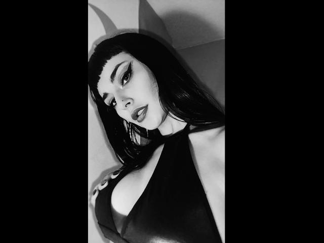 Black-and-white photo of Carll with long dark hair, heavy eyeliner, and full lips, wearing a halter top that reveals large, round tits. She's looking seductively at the camera.