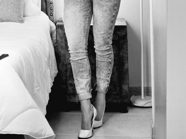 El trasero de Leared, vestido con unos vaqueros ajustados y con lazadas, está de pie, con tacones blancos, al lado de una cama con una colcha blanca y una mesita de noche de madera oscura, en una fotografía en blanco y negro..