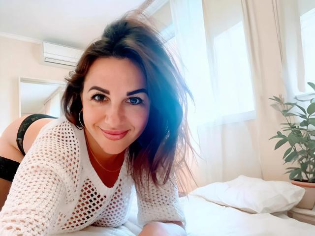 Dina69s is on a bed, smiling, wearing a white mesh top, black lace panties, and a necklace, with her tits slightly visible, brown hair, and a cute face.