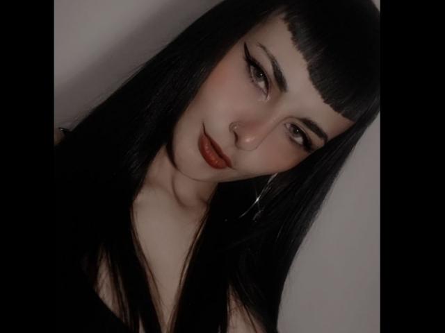 Carll has long black hair, straight bangs, dark eyeliner, red lipstick, and a nose ring. She's looking sideways with a slight smile, wearing a black top.