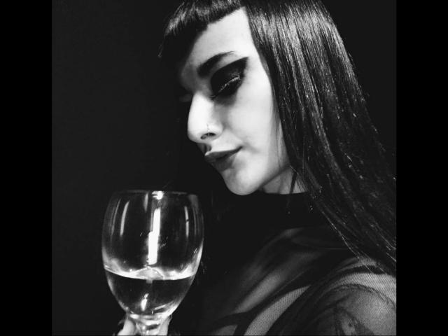 Carll with long dark hair, serious expression, holding a glass of water, wearing a sheer top, black and white photo, looking down, slight smile, glossy lips, dark background.