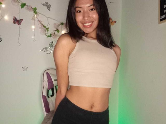 LuciaGarcia, with a cute smile, wearing a beige crop top and black pants, showing her flat stomach and small tits, standing in a room with butterfly decorations.