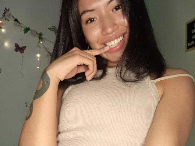 LuciaGarcia with dark hair, smiling, showing teeth, finger near mouth, wearing a beige ribbed tank top, tattoo on left arm, cute, attractive, brown eyes, light skin.