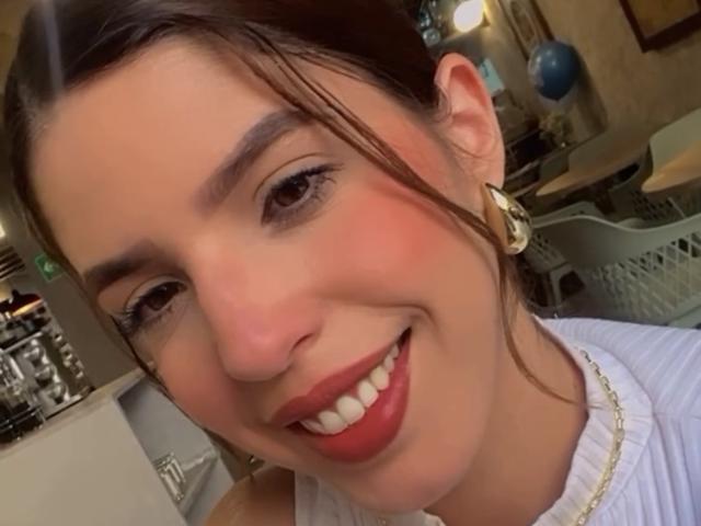 NatyBakers tiene ojos marrones, lápiz labial rojo y una sonrisa que muestra dientes blancos. Lleva una blusa blanca, aretes dorados y un collar. Su cabello castaño está parcialmente recogido..