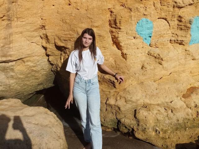 LauraLee69 with long brown hair, white t-shirt, and light blue jeans, standing against a rocky wall with a blue heart. Sun shadows on rocks. Hands relaxed, slight smile.