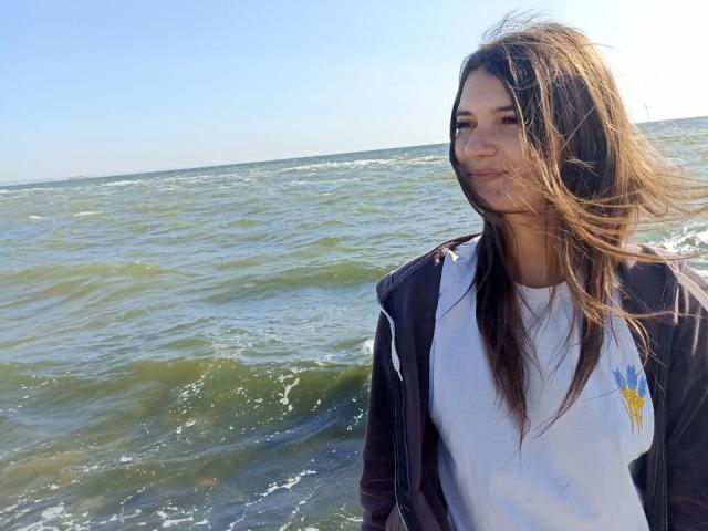 LauraLee69 with long brown hair, wearing a white shirt and black jacket, standing by greenish water, smiling slightly, wind blowing through her hair.