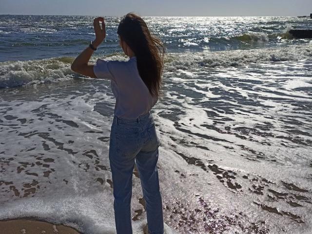 LauraLee69 stands at the beach with her back to the camera, wearing a white shirt and blue jeans, her brown hair in a ponytail, waving at the ocean.