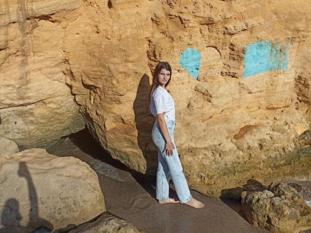 LauraLee69 stands barefoot against a rocky wall, wearing a white shirt and light blue, high-waisted, flared jeans, with her ass slightly pointed to the side.