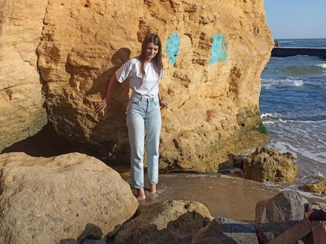 LauraLee69 stands barefoot on rocky beach, wearing a white shirt and high-waisted light blue jeans, leaning against a large, textured yellow rock with blue graffiti.
