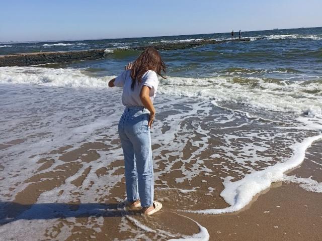 LauraLee69 stands barefoot on a sandy beach, facing the ocean, wearing a white shirt and high-waisted blue jeans, with her brown hair down, waves touching her ass.